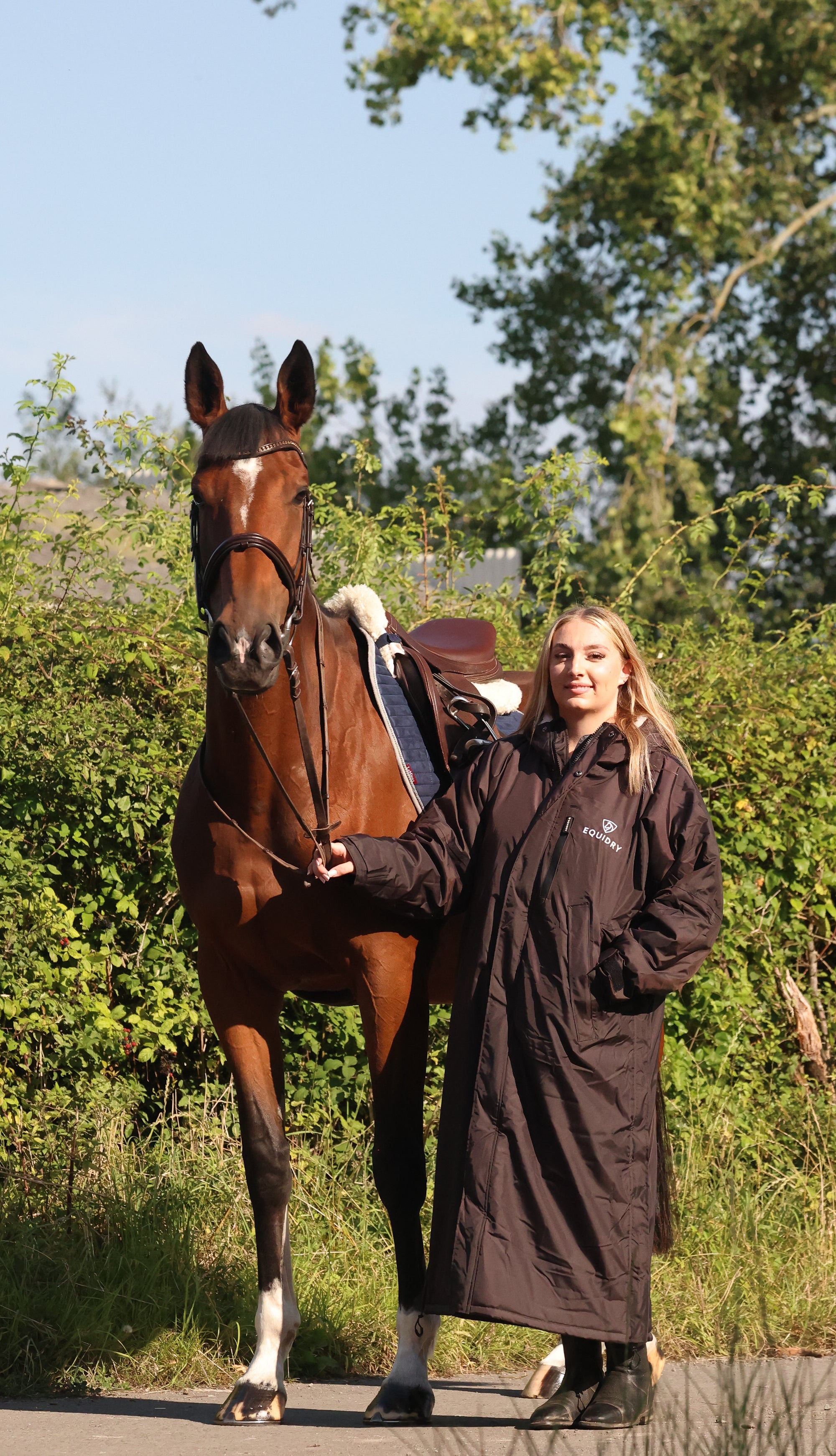 EQUIDRY women's long waterproof horse riding coat in rich brown/cream modelled by rider stood with horse