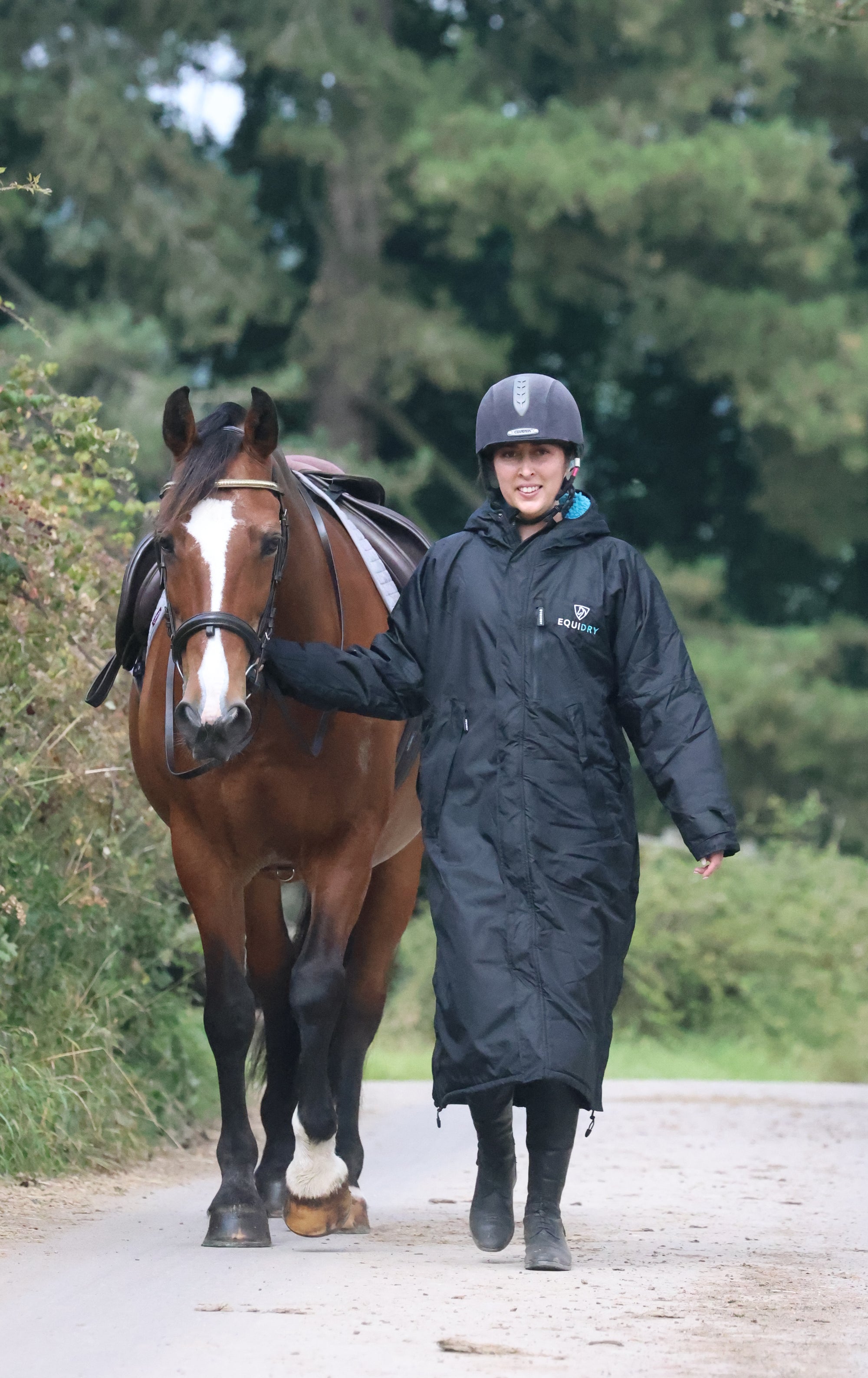 EQUIDRY women's long waterproof horse riding coat in black/turquoise modelled by rider walking with horse 